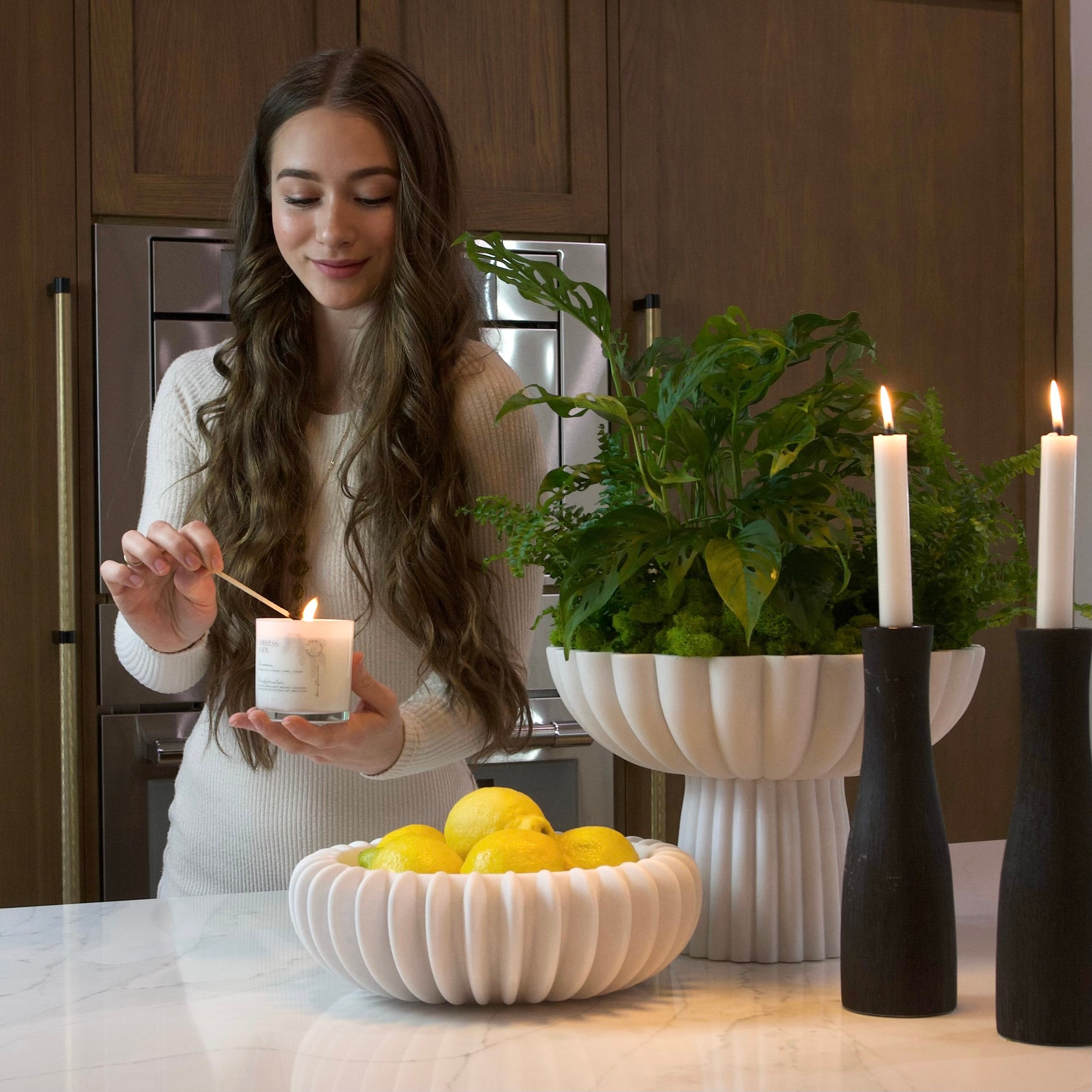 Woman lighting a candle in a kitchen with a bowl of lemons and a plant.